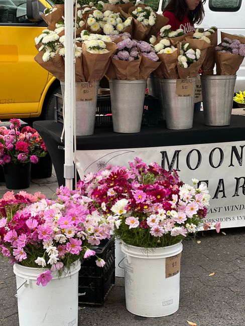 A floral display featuring multiple bouquets of fresh flowers arranged in brown paper wrapping, placed in metallic buckets on a black table, with a mix of white, pale purple, and cream blooms. In fron