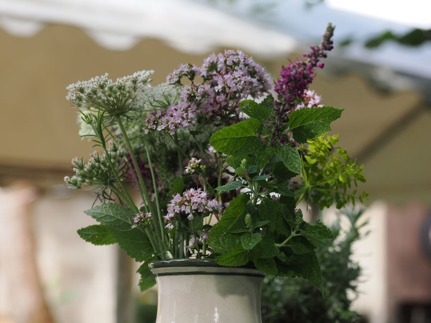 A close-up of a floral arrangement in a white ceramic vase, featuring fresh herbs and flowers including mint with textured green leaves, pink and purple lavender with slender spiked stems, and cluster