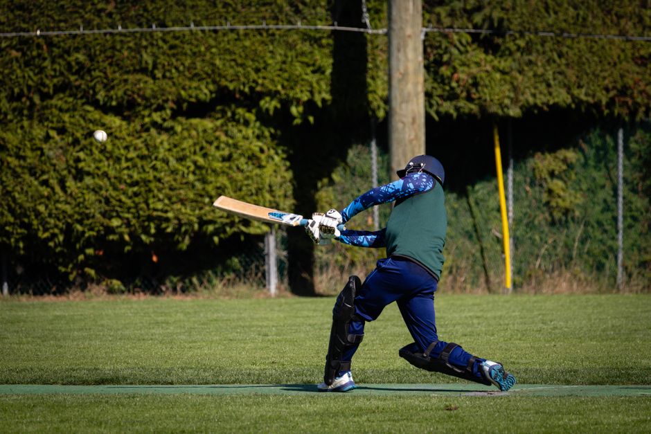 A cricketer in protective gear, including a helmet, pads, and gloves, is captured mid-swing on a grassy field during daytime, preparing to hit a white cricket ball with a wooden bat. The player is wea