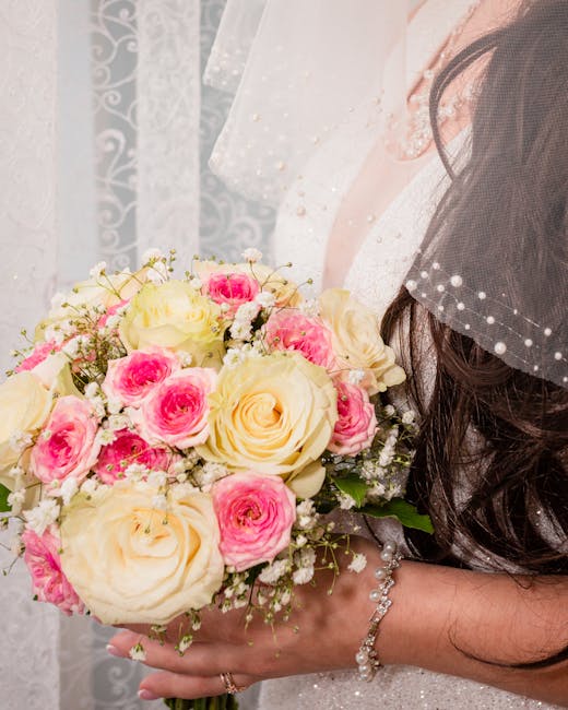 A bride holding a bouquet arrangement of fresh flowers featuring large cream roses and smaller pink roses, accented with delicate white baby's breath, wrapped in light pink tissue paper and tied with